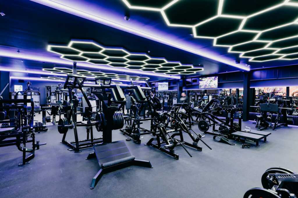 Fitness Cartel Interior; A well-lit gym interior with various exercise equipment arranged throughout the space, including treadmills, weight benches, and elliptical machines. A purple light fixture hangs from the ceiling.