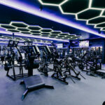 Fitness Cartel Interior; A well-lit gym interior with various exercise equipment arranged throughout the space, including treadmills, weight benches, and elliptical machines. A purple light fixture hangs from the ceiling.